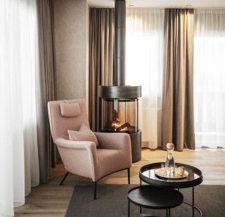 Peternhof_Suite_Leseecke mit Kamin.jpg Leseecke mit Fauteuil in Rosè, dahinter Feuerstelle, davor zwei Beistelltische, Deckenausschnitt, Vorhänge in zwei Farben